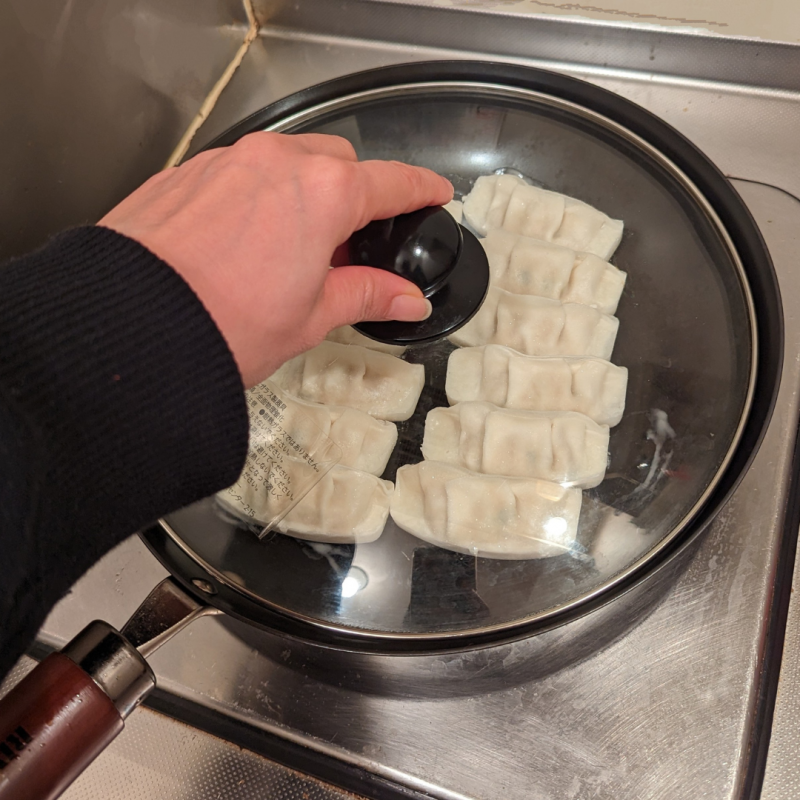 キッチンのひとくちコンロにリバーライト極JAPANフライパンをかけ、餃子を焼いている画像。餃子をフライパンに6個ずつ2列に並べガラスの鍋フタをして蒸し焼きにする様子。鍋フタのツマミを左手でつまんでいる。