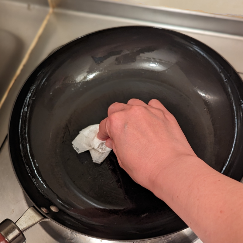 キッチンのひとくちコンロにリバーライト極JAPANフライパンをかけ、右手でキッチンペーパーを使ってフライパンの　鍋肌に油を擦り込んでいる。