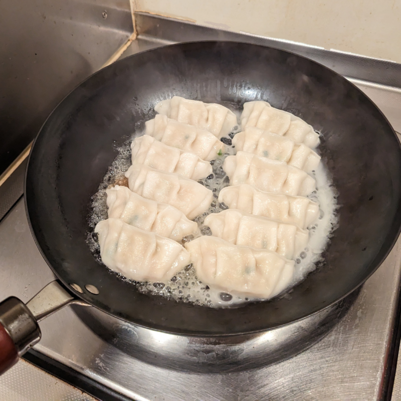 キッチンのひとくちコンロにリバーライト極JAPANフライパンをかけ、餃子を焼いている画像。

鍋フタを取り、火を強めて水分を飛ばしながら焼いている様子。餃子の周りで水分がジュウジュウと蒸発しているのがわかる画像。